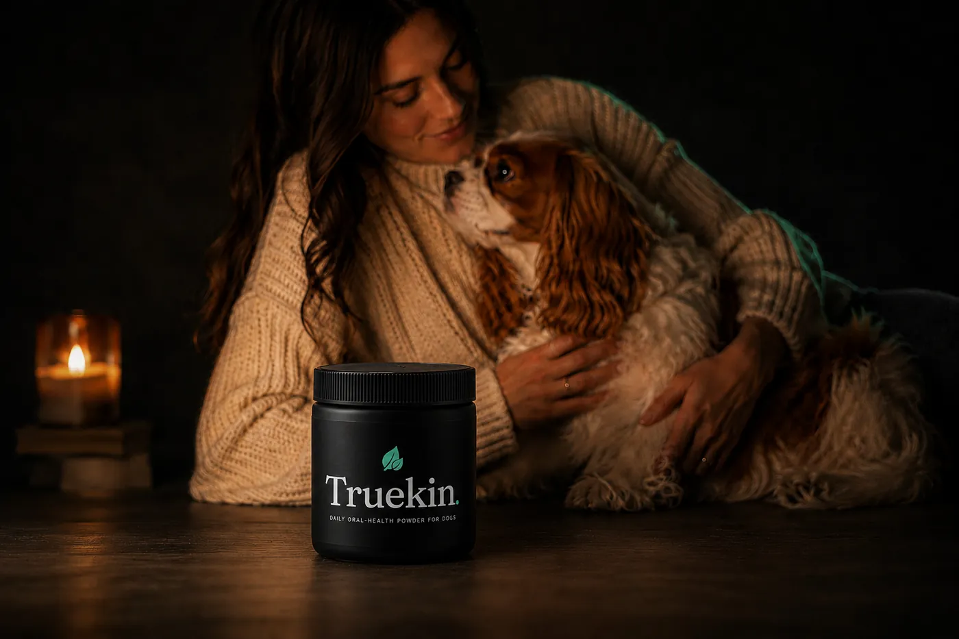Owner sitting on a dark floor with her Cavalier King Charles Spaniel, a jar of Truekin daily oral-health powder in the foreground